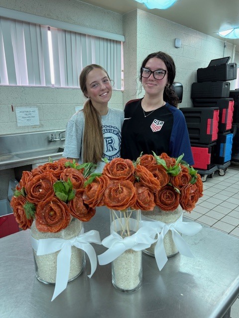 Two women stand next to a table holding three vases filled with orange, rose-shaped items.
