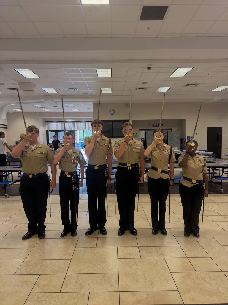 Five uniformed individuals stand in a row holding swords, with two others in the background. They are indoors in a tiled room.