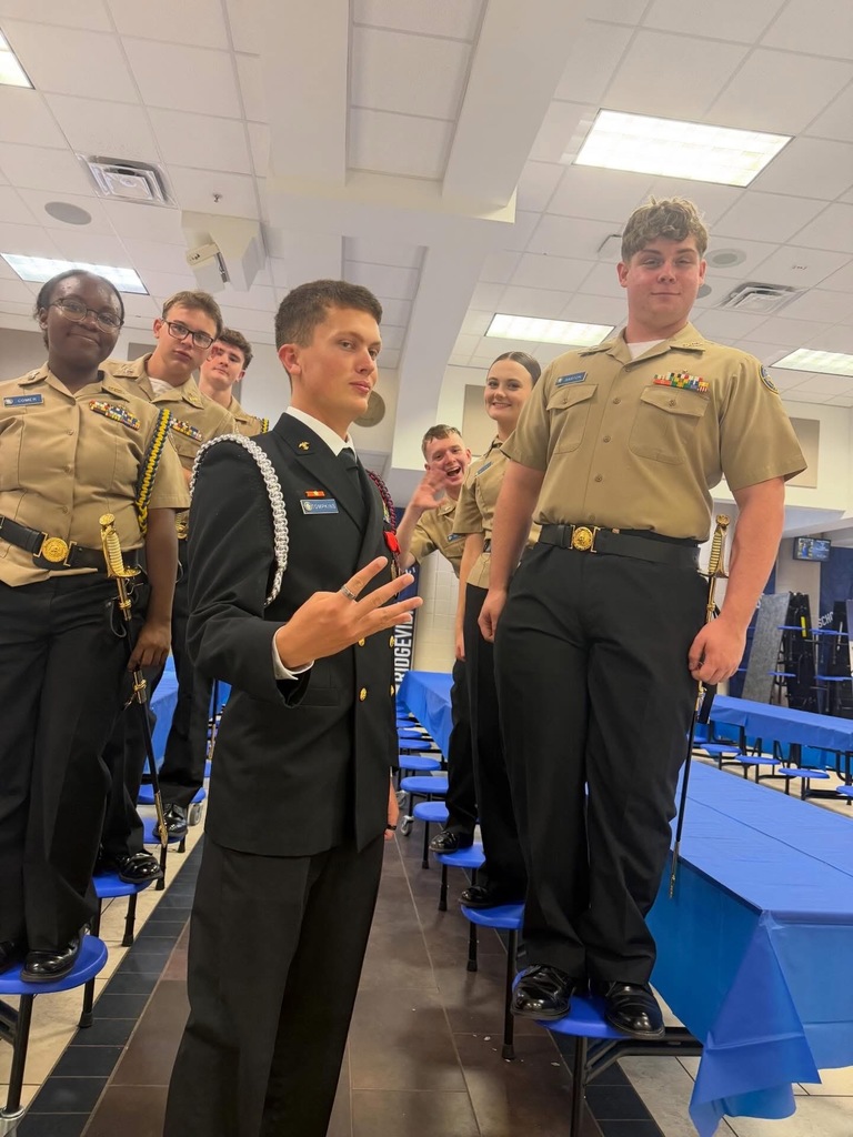 Several people in uniform, including one with a hat, are standing in a room with blue tables.