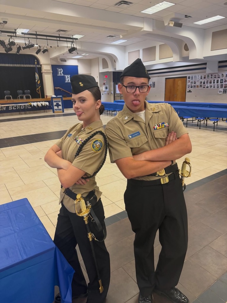 Two uniformed individuals, one female and one male, stand with arms crossed in a room. Behind them is a table draped in blue fabric.