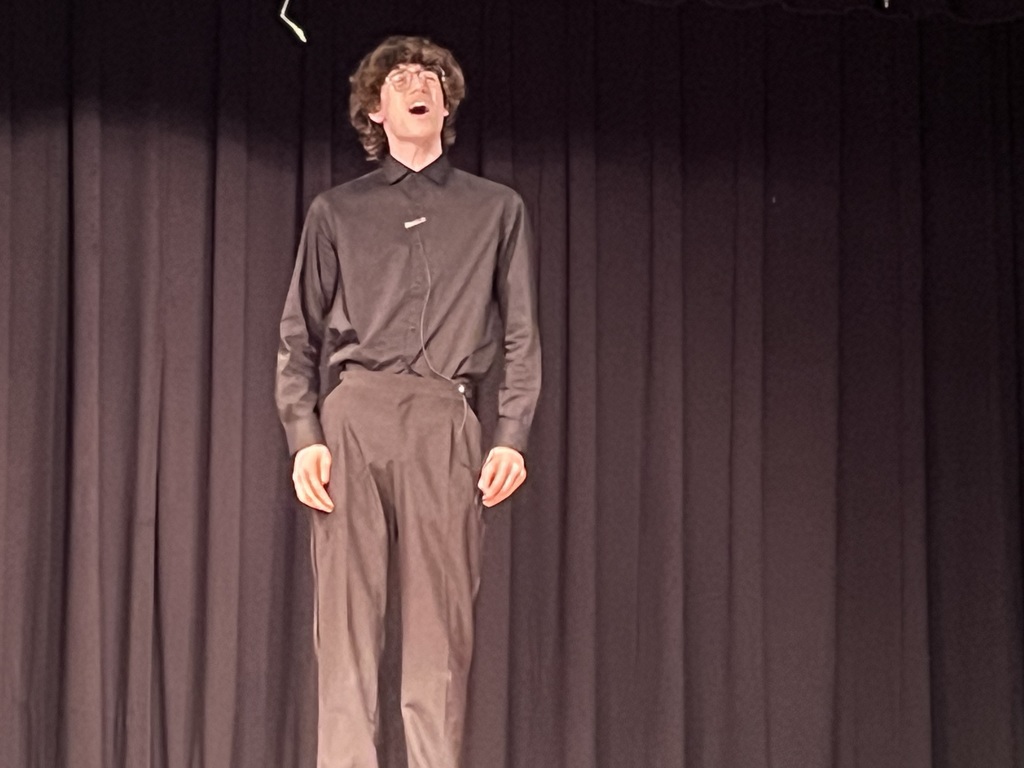 A person stands on stage, singing, dressed in a black shirt and gray pants, against a black curtain backdrop.