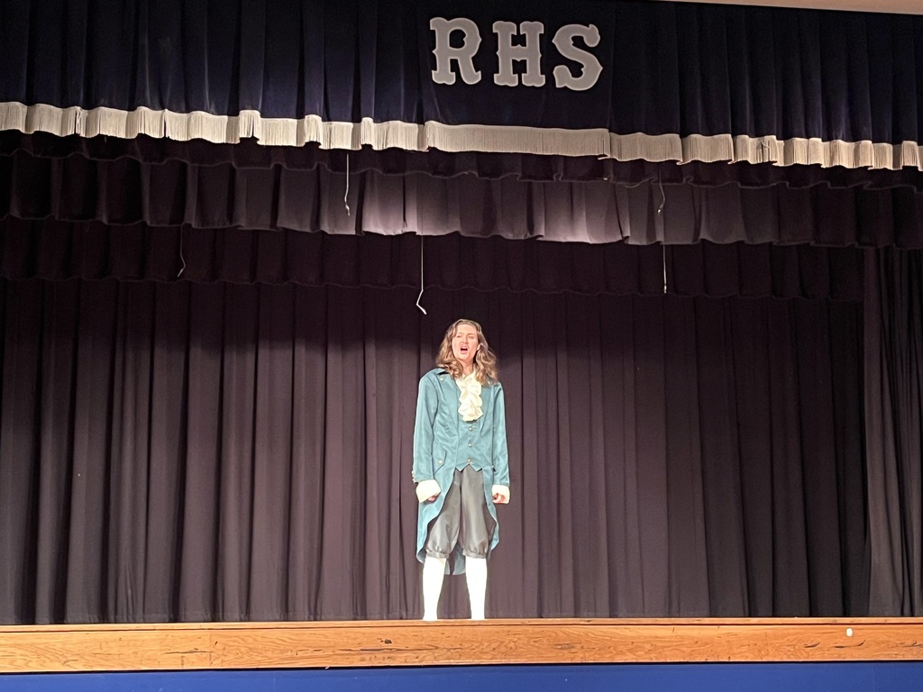 A woman in a blue coat stands on a stage with a black curtain and wooden floor.