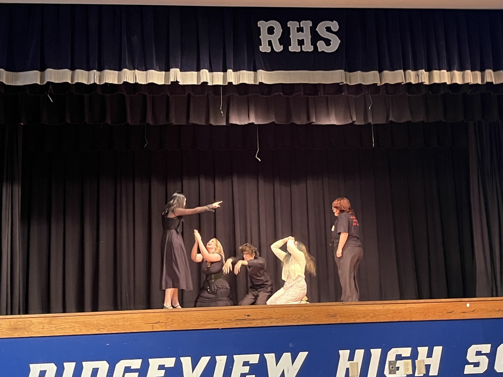 Five individuals on stage, four in black and one in white, against a blue backdrop with "RIDGEVIEW HIGH SCHOOL" sign.