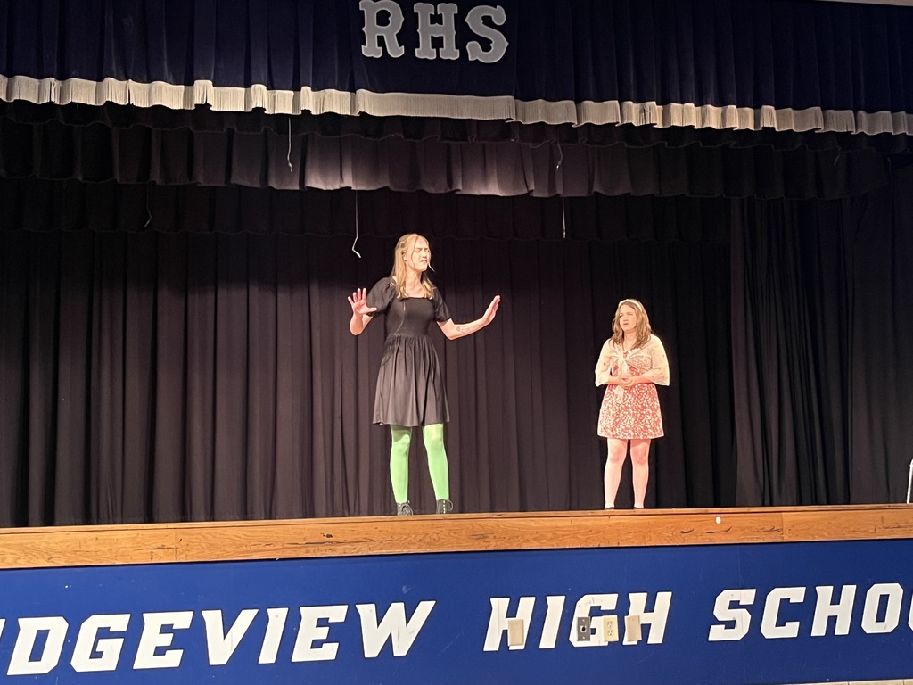 Two women stand on a stage, one in a black dress and green socks, the other in a pink dress.