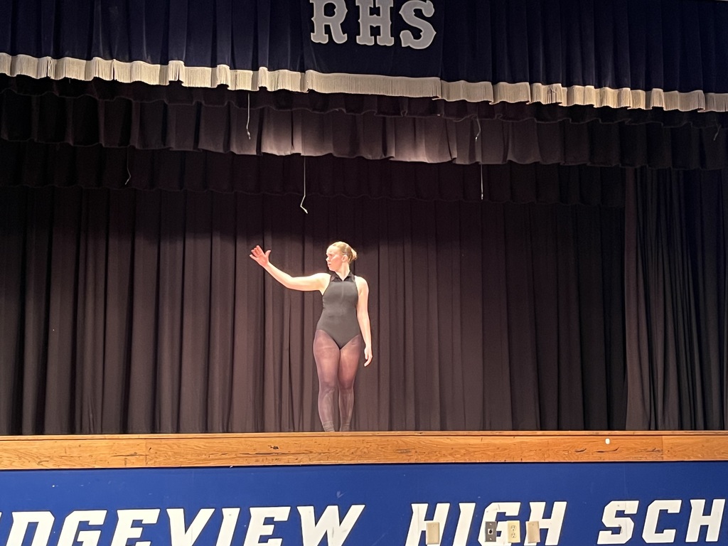 A person in a black leotard stands on a stage at Ridgeview High School, with arms raised.