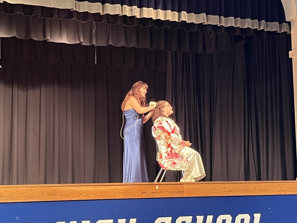 A woman on stage styles another woman's hair. Black curtains and a blue backdrop are in the background.