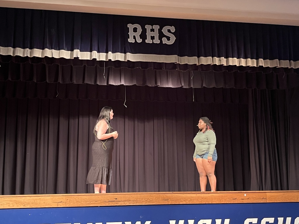 Two women stand on stage. One wears a dress; the other, a green top and shorts. Behind them, a black curtain displays "RHS".