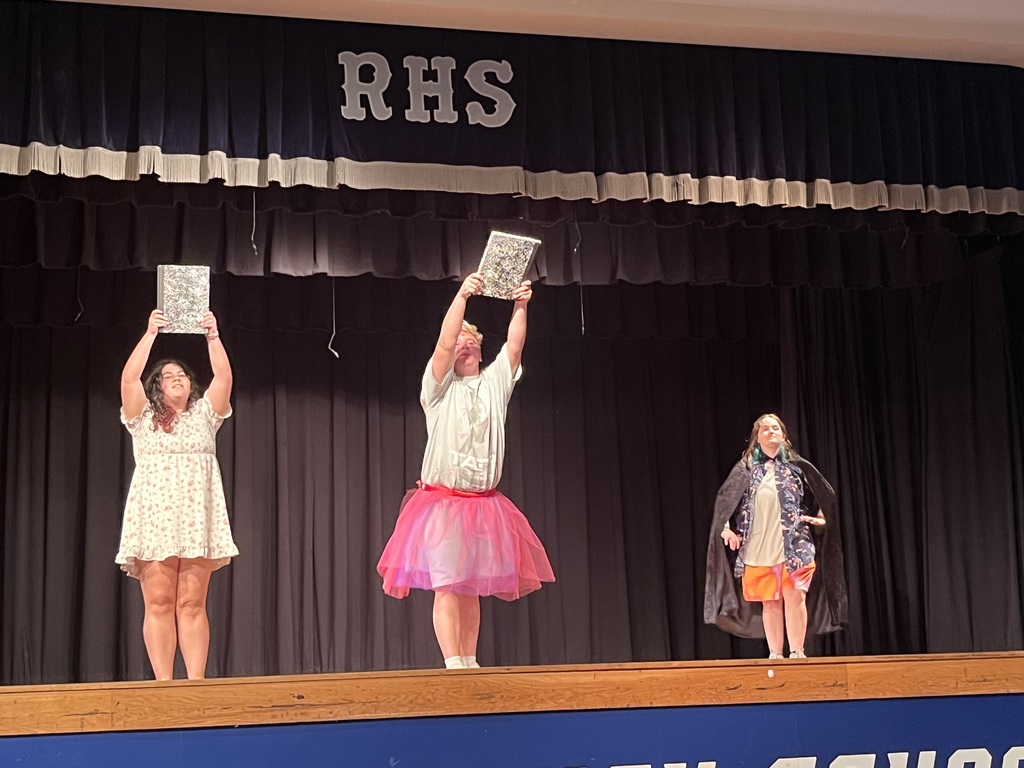 Three individuals on stage, holding signs, in front of a black curtain with "RHS" on it.