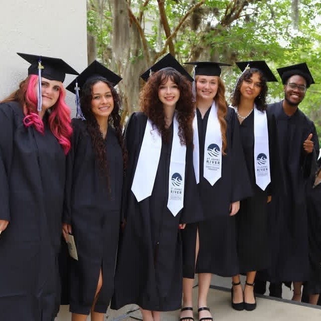 A group of people in graduation robes, some with sashes, smiling in front of trees.
