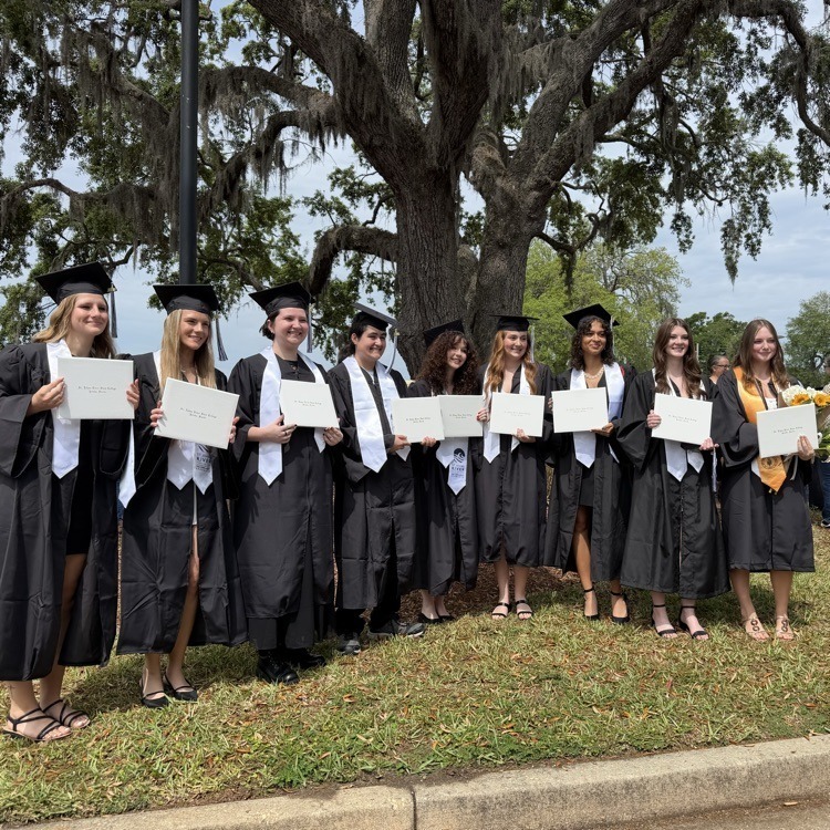 Graduates in black gowns and caps stand in a row, holding diplomas, on grass near a tree.