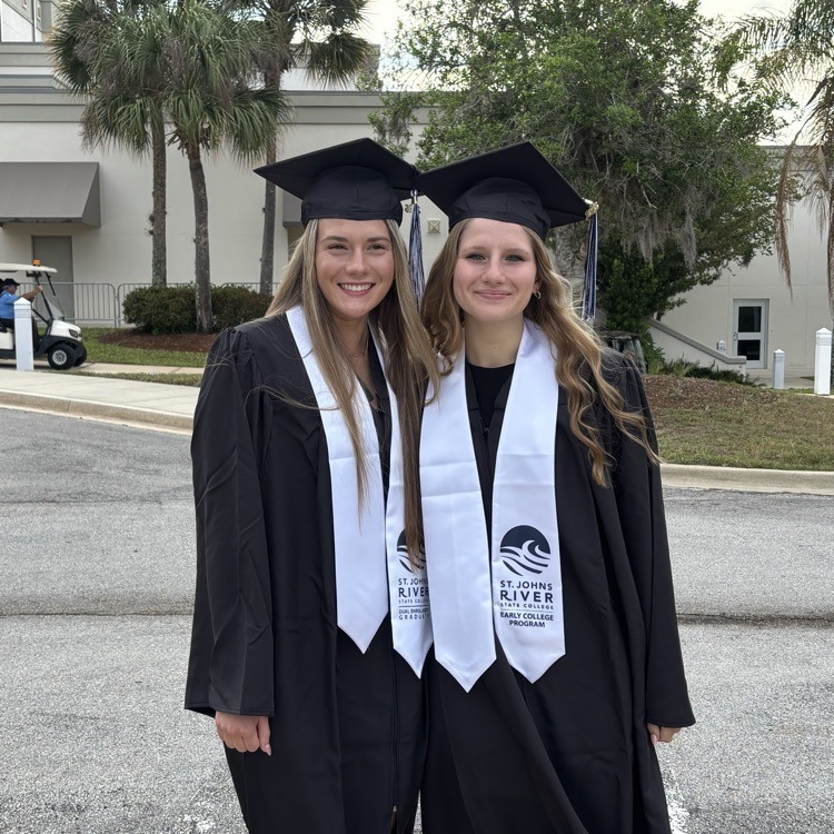 Two women in graduation gowns, smiling and standing next to each other, with a building and trees in the background.