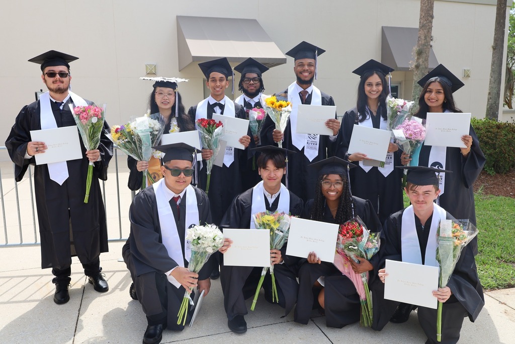 Group of students in graduation robes holding certificates and flowers, standing and sitting in front of a building.