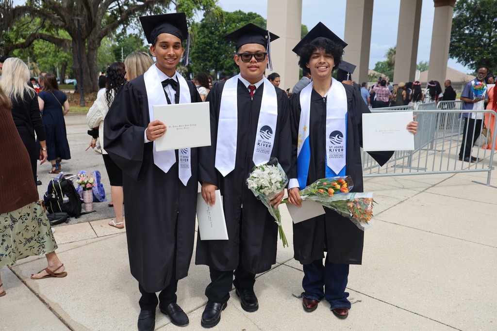 Three men in graduation gowns and hats stand together, holding diplomas and flowers, in front of a crowd.