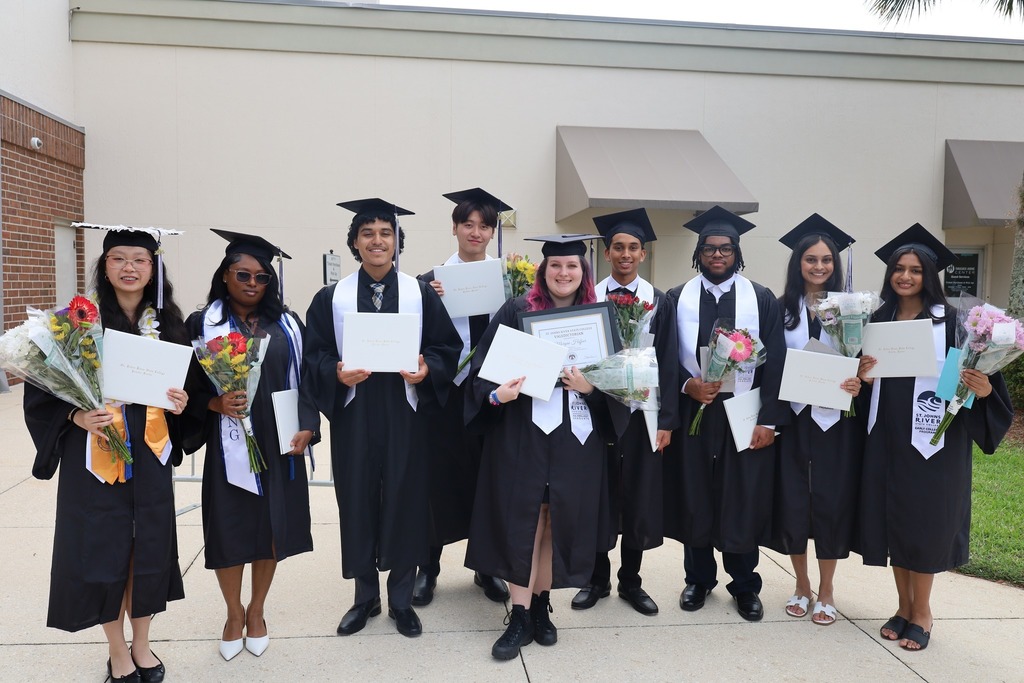 A group of graduates in caps and gowns hold certificates and flowers in front of a building.