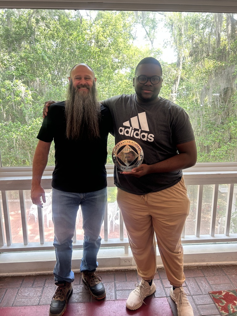 Two men stand on a porch. One wears a long beard and holds a trophy. Both wear sneakers.