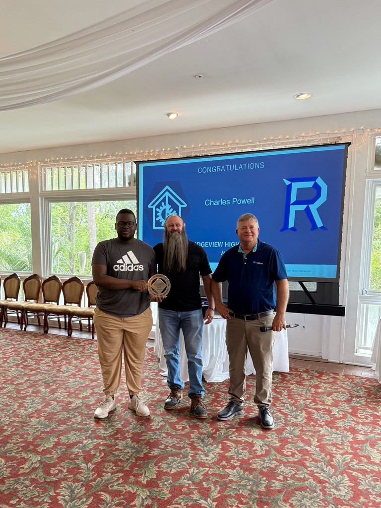 Three men stand in front of a projector screen. One man holds a trophy. They wear casual attire. A carpeted room with chairs and windows is in the background.