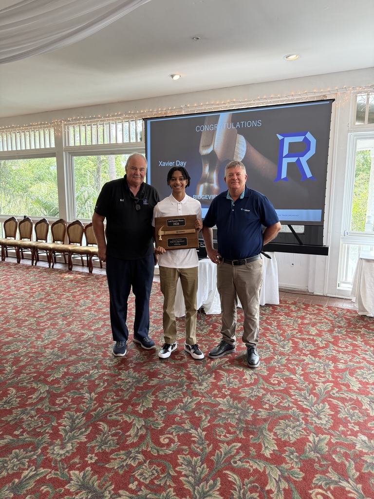 Three people stand in a room with a projector screen displaying an "R" logo. One holds a trophy.