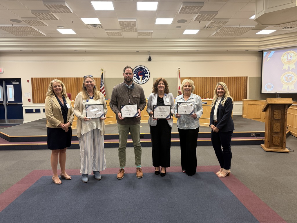  An award presentation at a Clay County event. Six people are standing in a line on a stage in front of a wood-paneled wall with the Clay County seal. Four people in the middle are holding framed certificates. The two people on the ends are standing with their hands clasped. They are all smiling at the camera. The room has a blue carpet and a large screen on the right side.