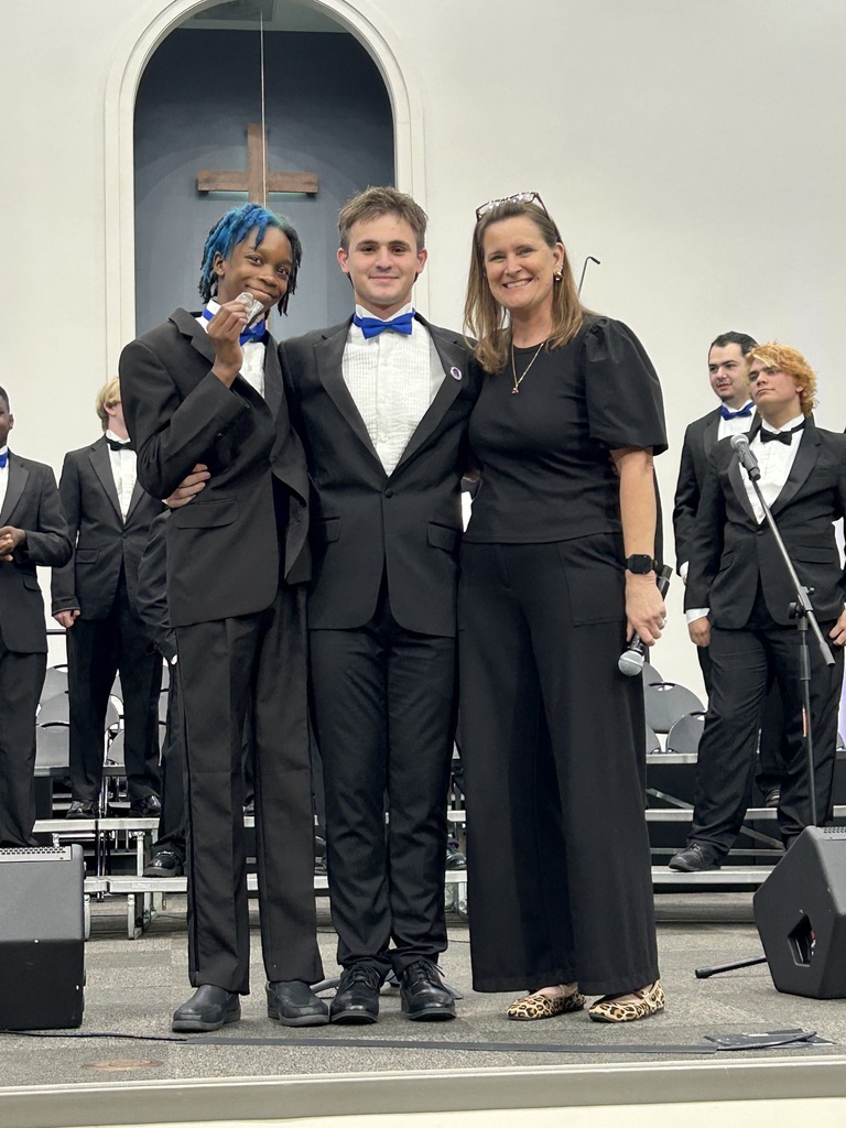 Three people stand on a stage. A man on the left holds a microphone. Behind them, musicians in black suits and white shirts perform on stage.