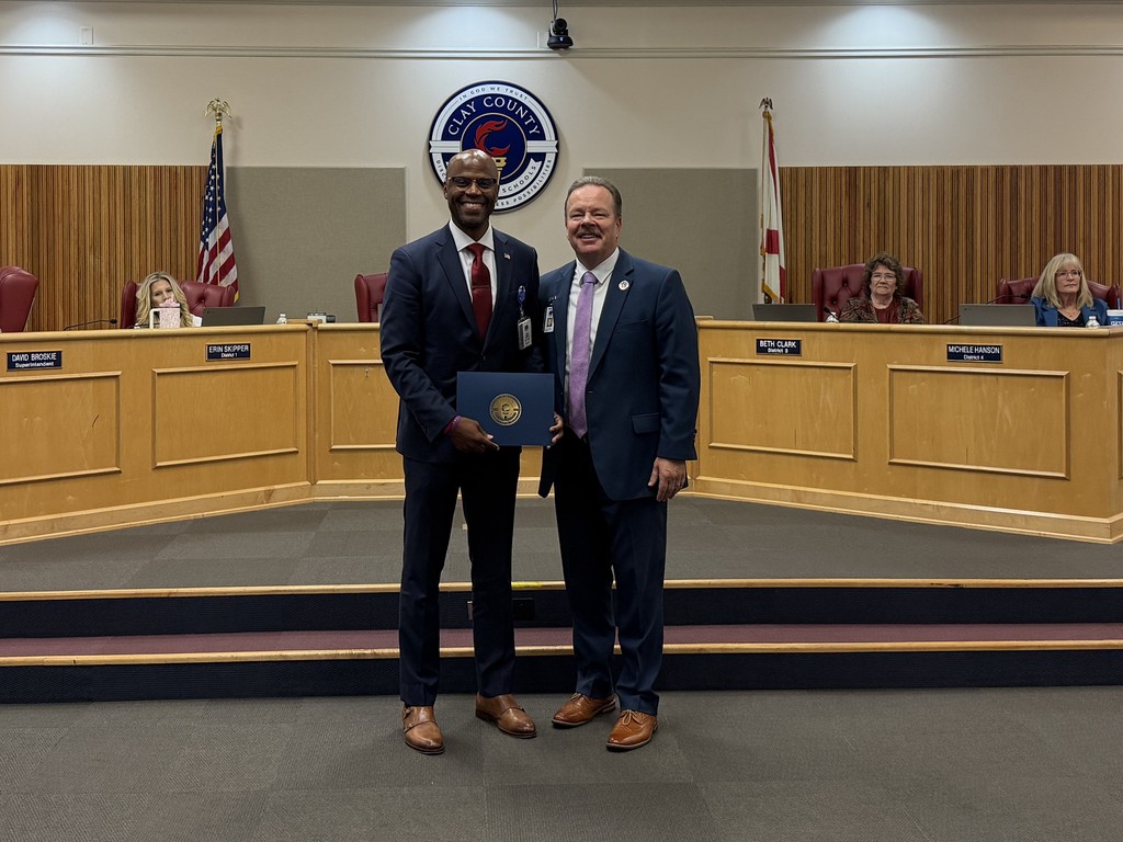 Two men in suits and ties stand in a board chamber, one holding a certificate. Behind them, flags and wooden benches.
