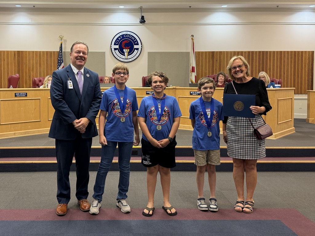 A group of five stands in a room, with four young boys wearing blue shirts and medals, and a woman holding a certificate.