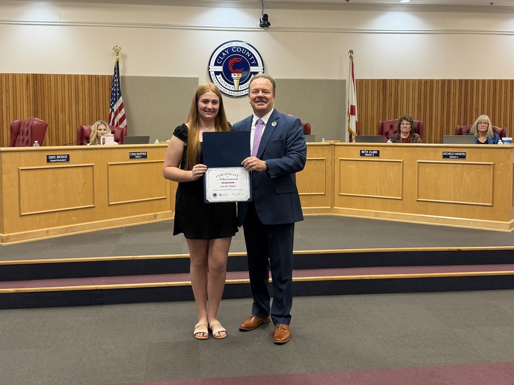 A man and woman stand on a stage in a board room holding a certificate. Behind them is a podium.