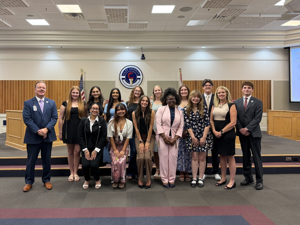 Group of students and faculty in formal attire standing together on a stage in a board room.