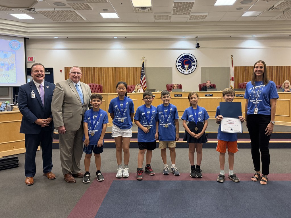 Students in blue shirts, some with medals, stand with adults in formal wear in a meeting room.