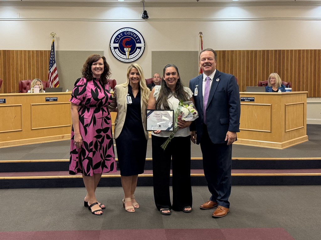 Four people stand in a board room. A woman holds a framed award and flowers. 