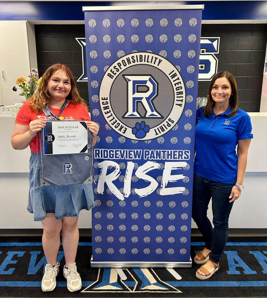 Two women stand next to a banner reading "RISE" with the "R" logo, one holding a certificate.