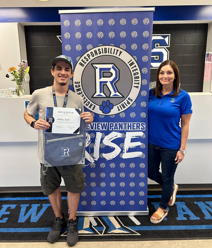 Person in a cap stands next to a blue banner that reads "RISE" in front of a white wall.