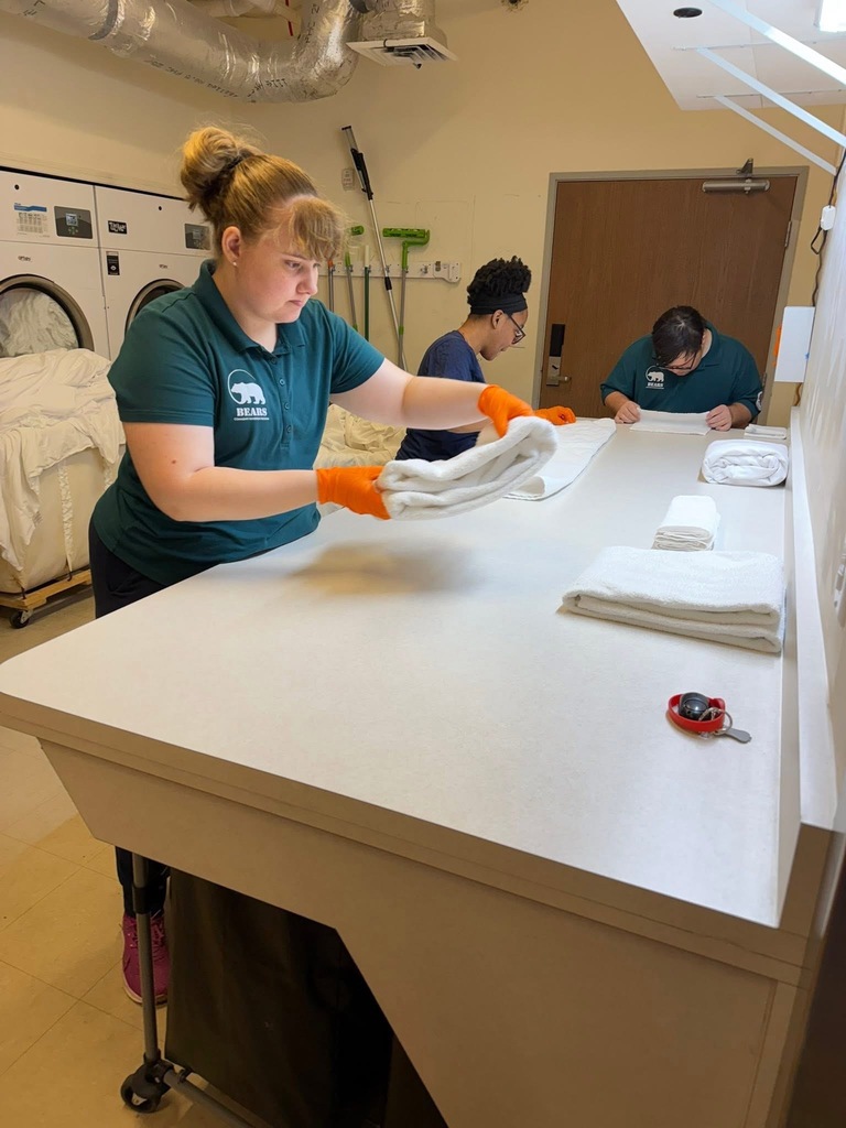 A person wearing a green shirt and orange gloves folds towels. Two others are working in the background.