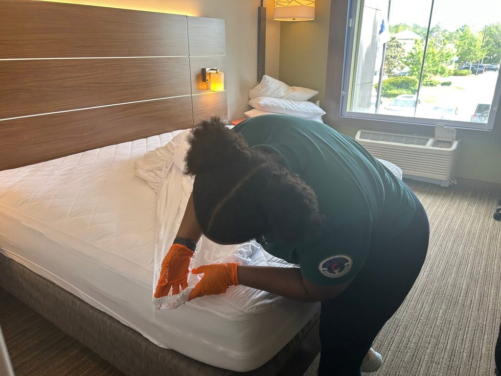 A person in green scrubs and orange gloves adjusts a bedcover on a bed in a room.