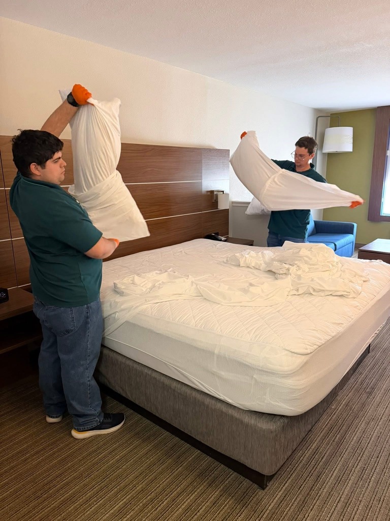 Two people work on a bed, holding pillows, in a room with a blue chair.