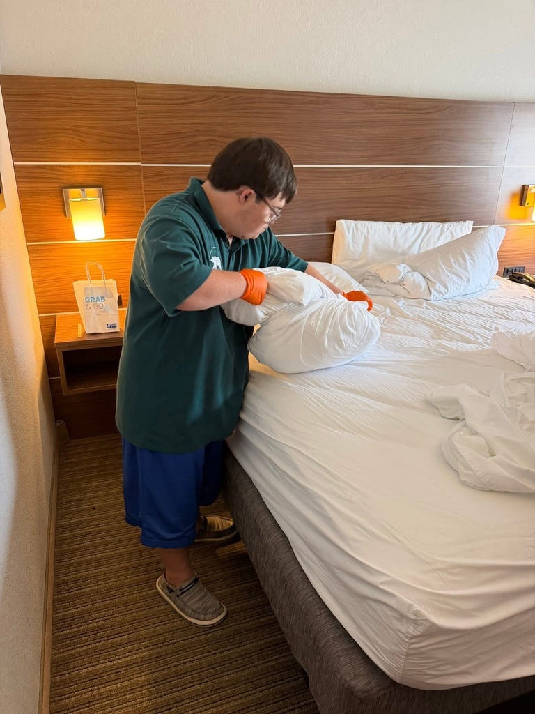 A person in a green shirt and blue shorts adjusts a pillow on a hotel bed.