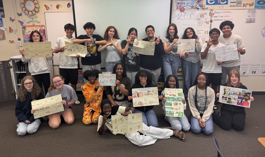 A group of people in a classroom holding signs, some sitting and others standing, with a whiteboard and bulletin board behind them.