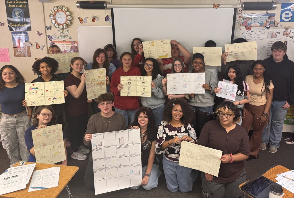 A classroom full of students posing with their artwork, smiling, and holding up pieces of paper.
