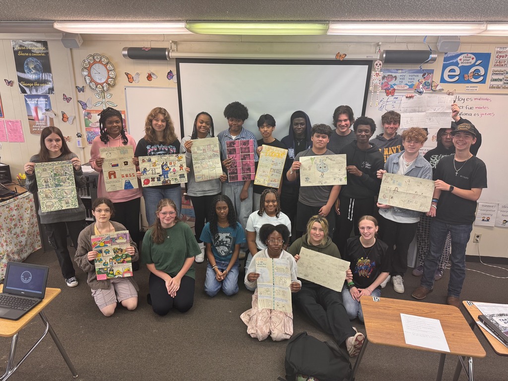 A group of students in a classroom holds up drawings while sitting and standing in front of desks.