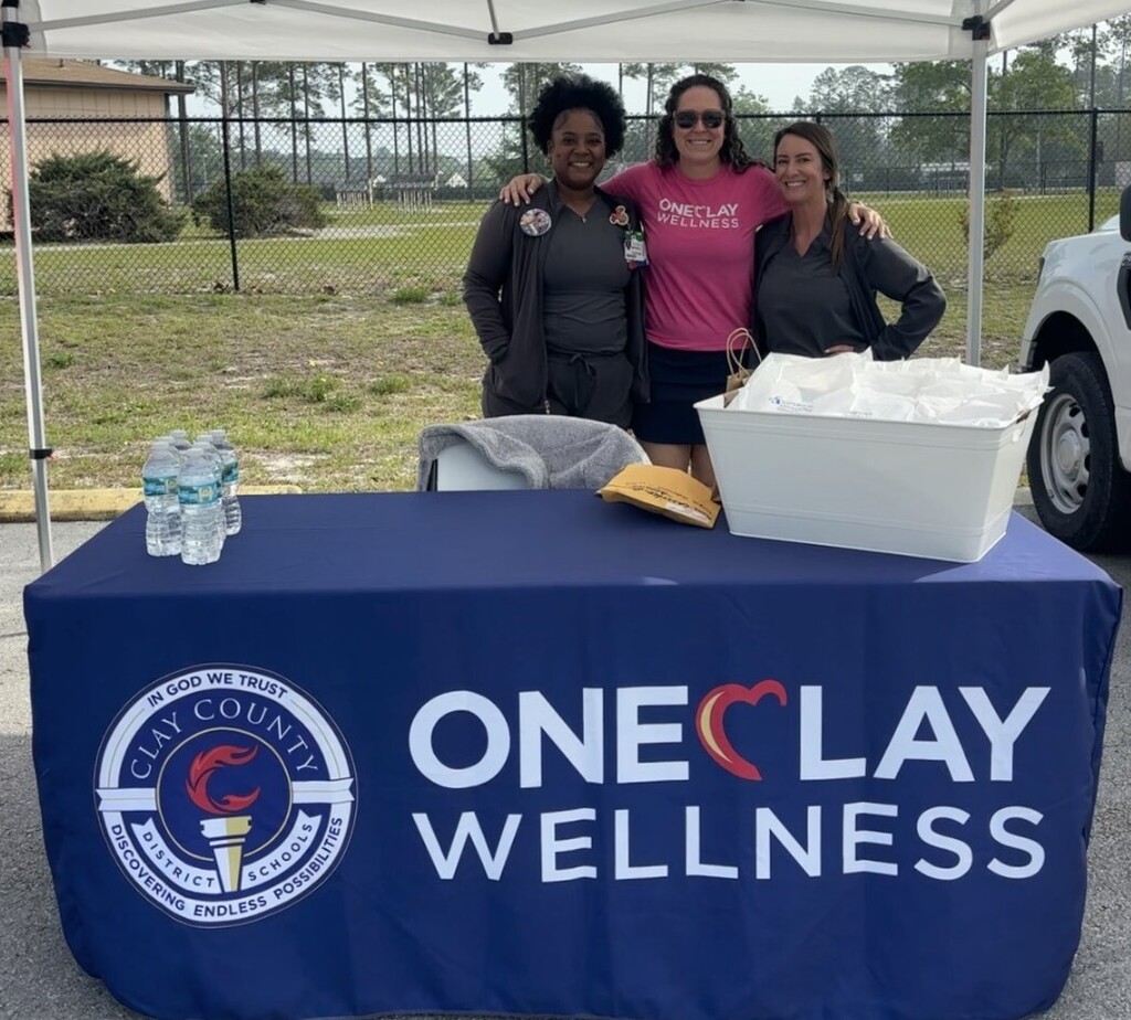 Three people stand behind a table with a "One Clay Wellness" banner and  a tent