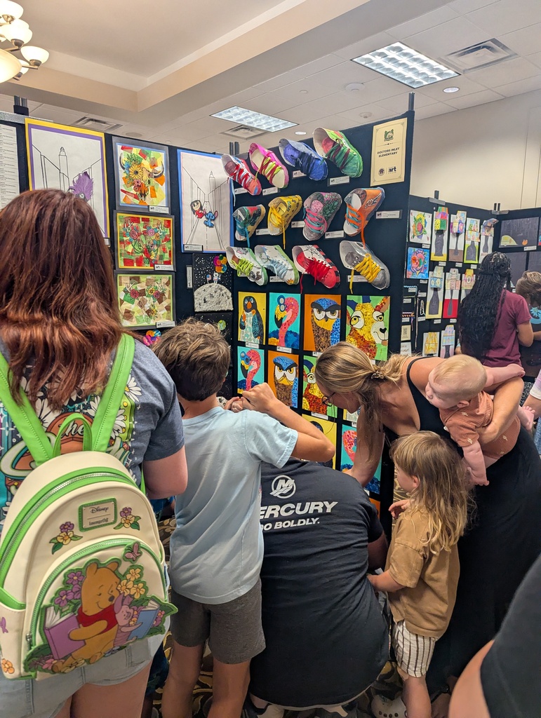 Group of people in a room, standing around and looking at a display of artwork on a wall.