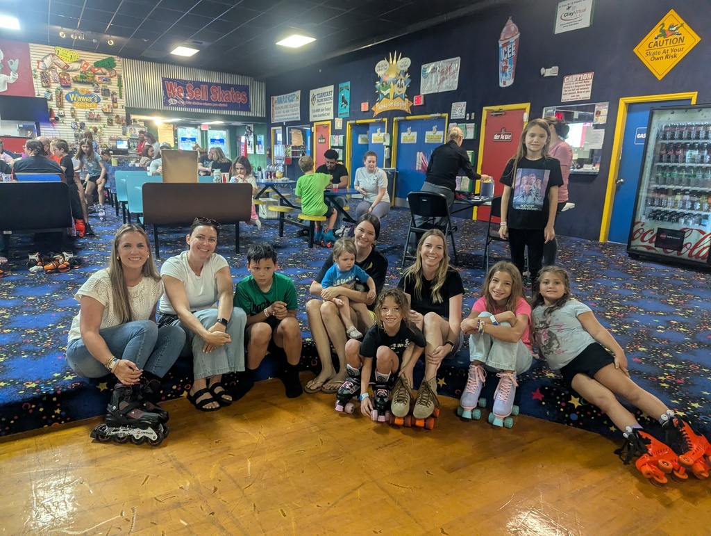 A group of people in a roller rink, sitting and standing with roller skates on, surrounded by a colorful, playful wall.
