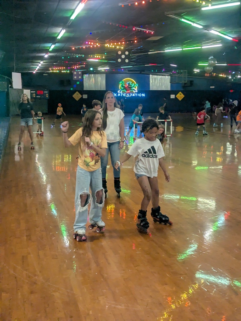 A roller rink with children and adults wearing skates. A woman assists two children, one holding hands.