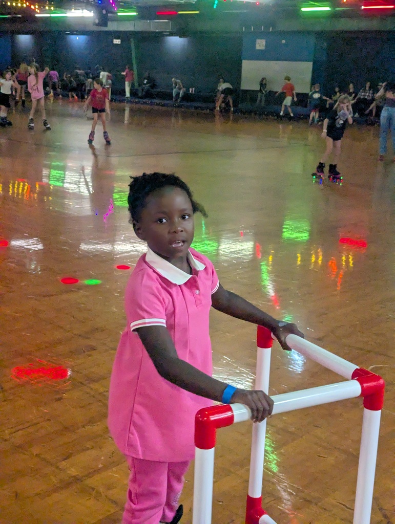 A young girl stands by a red and white railing in a roller rink. Other children skate around her.