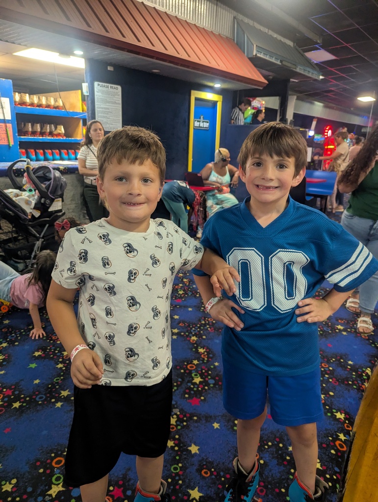 Two boys stand in an indoor area with multicolored flooring. One wears a white T-shirt with black patterns, and the other wears a blue jersey with "00".