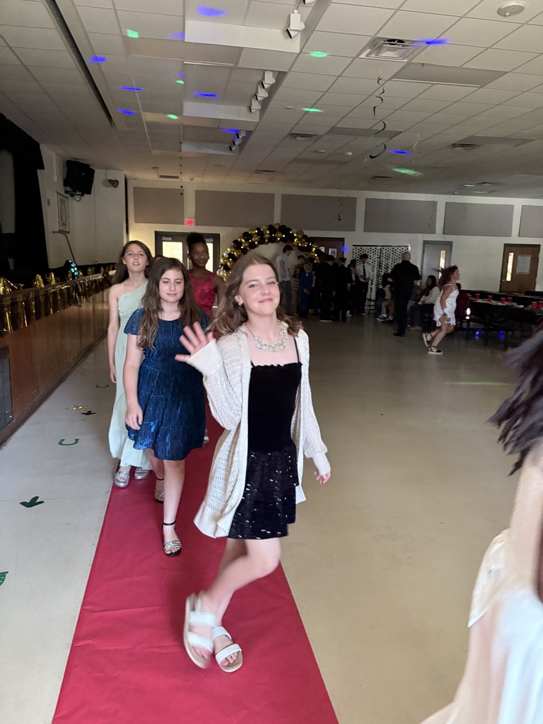 Several young women walk on a red carpet inside a hall. Some people are standing behind them.
