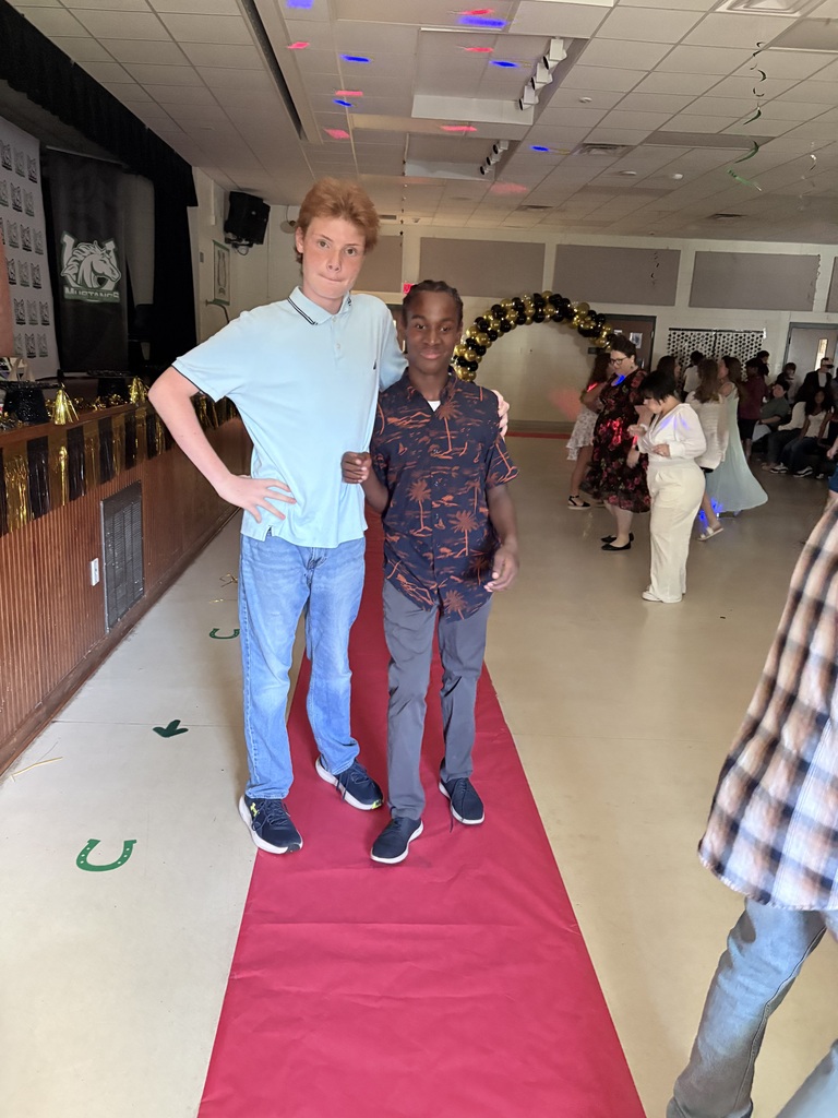 Two boys stand on a red carpet; one in a blue shirt and the other in a patterned shirt.