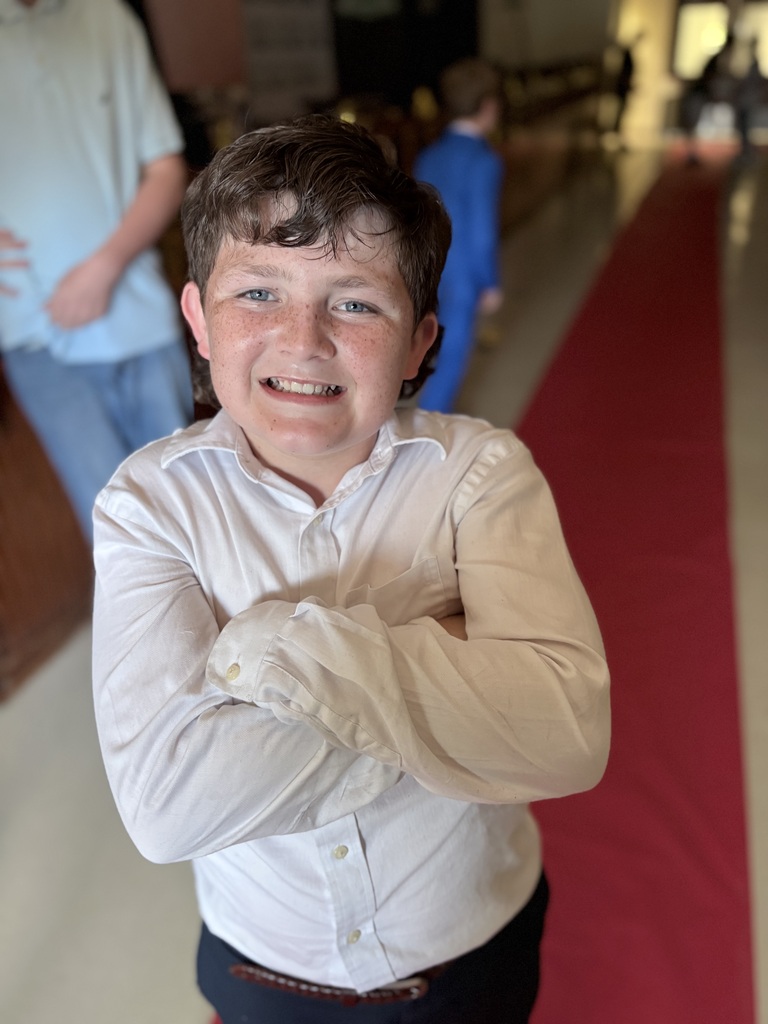 A smiling boy with folded arms in a white shirt stands on a red carpet in a corridor.