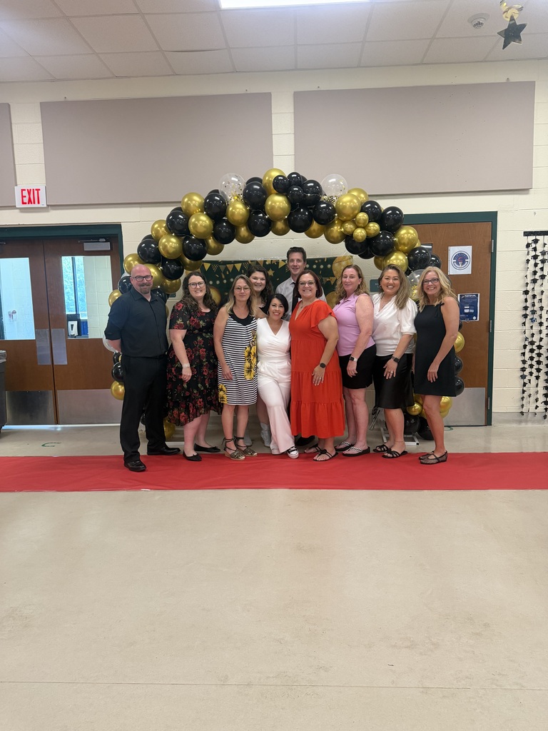 A group of people in a hallway. Some wear dresses, others wear suits. They stand under a gold and black balloon arch.