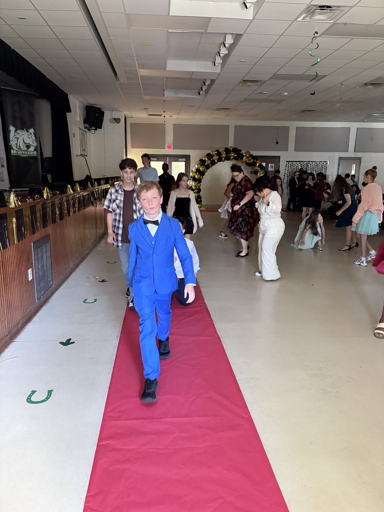 A boy in a blue suit walks on a red carpet inside a room, while others move around.