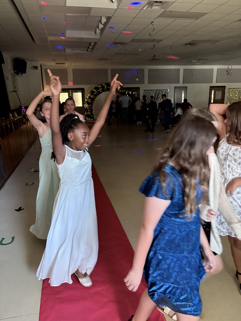 Several children in various dresses walk on a red carpet at an indoor event.
